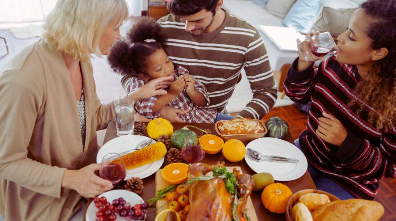 A family sharing a warm meal together during an autism-friendly Thanksgiving, with adults gently supporting a young child at a festive table filled with turkey, autumn produce, and sensory-friendly holiday foods.