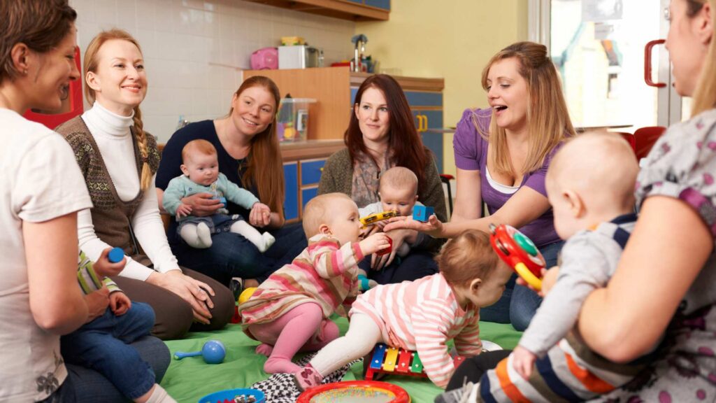 Group of mothers with their babies sit together on a playmat sharing conversation and toys, representing an autism parent support groups