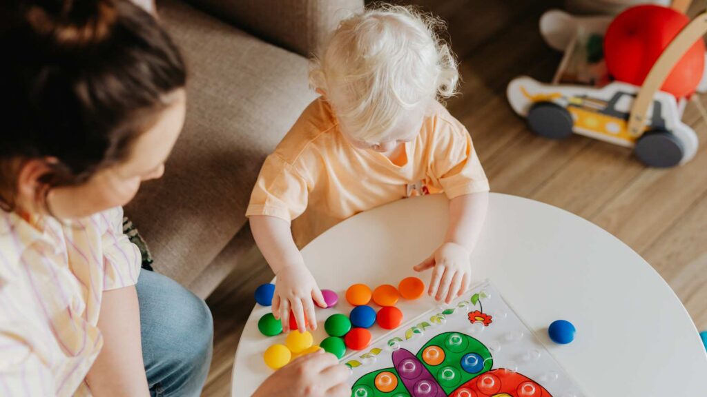 Toddler sorts colorful pegs into a shape board while a caregiver observes, representing how early should ABA therapy start through play-based learning activities