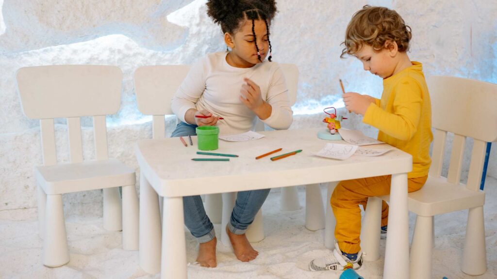 wo young children sit together at a small white table drawing and playing with toys, illustrating making friends with autism through shared play activities